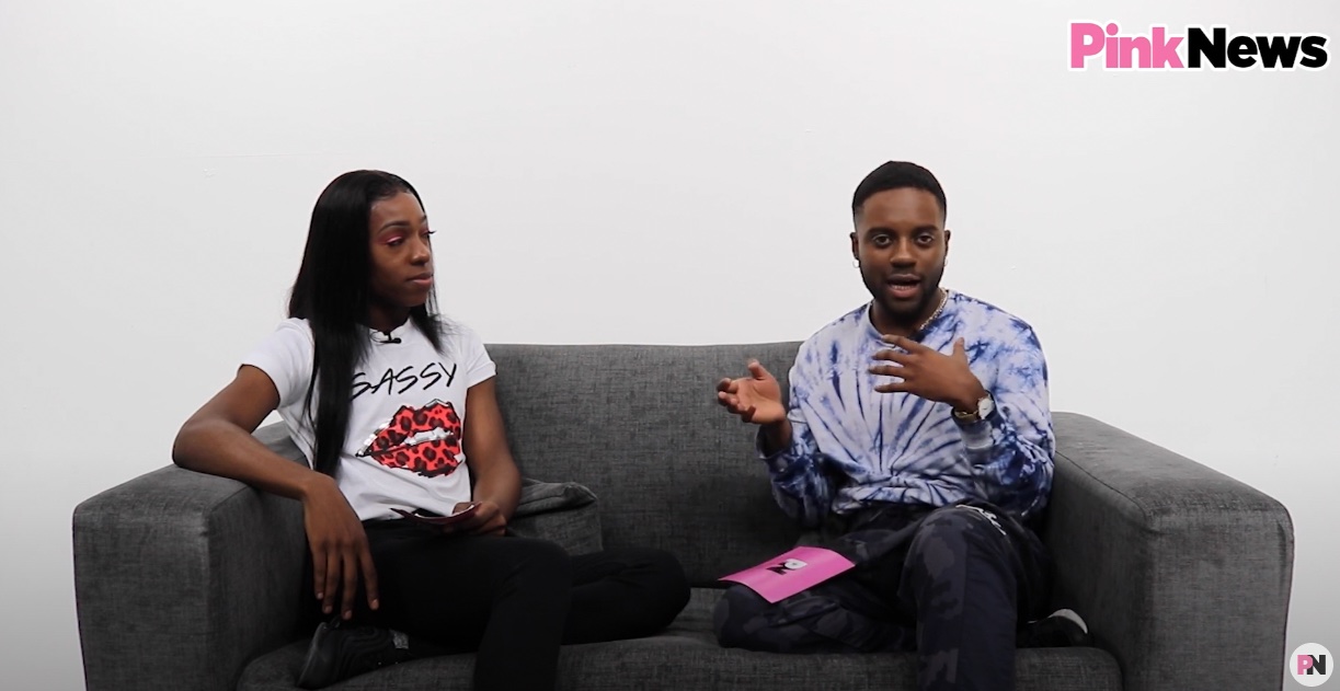 This is an image of two Black men. On the left, he is wearing a white tshirt and has long black hair. On the right, he is wearing a tyedye shirt. They are both sitting on a grey couch in a white room.
