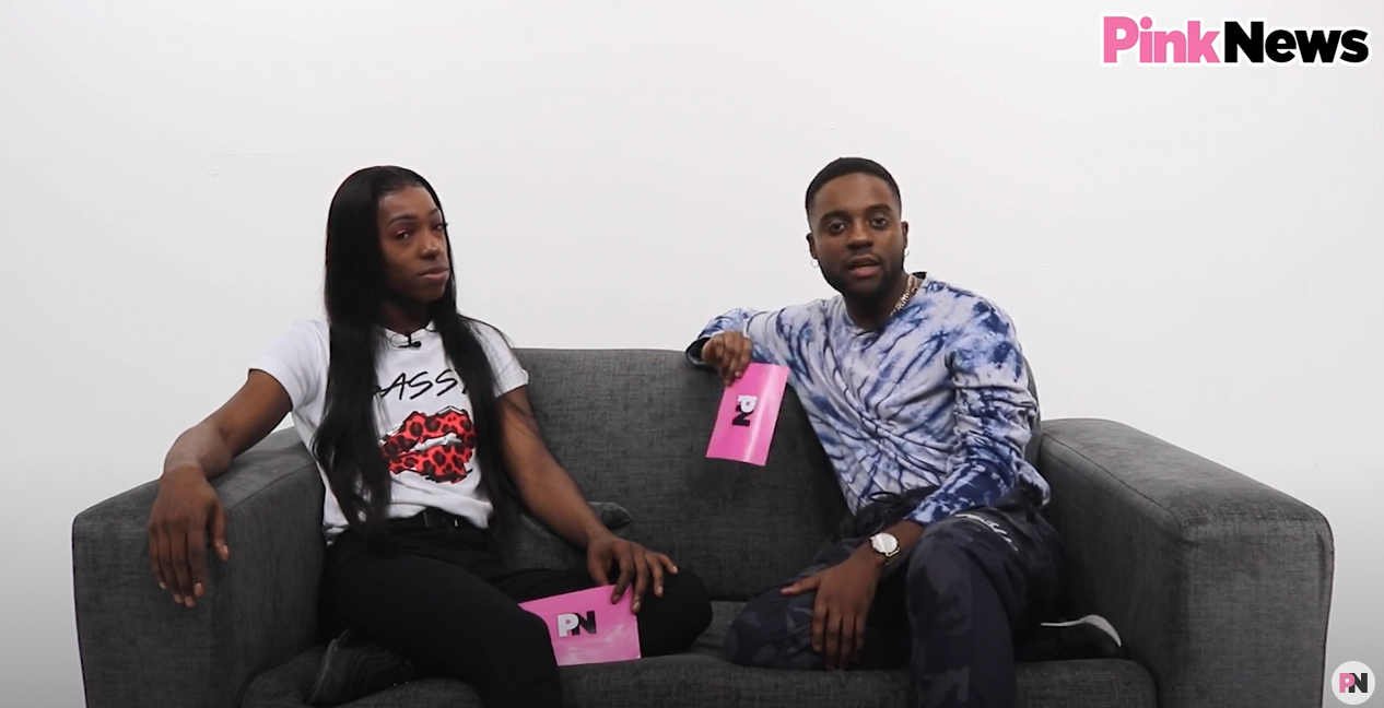 This is an image of two Black men sitting on a couch in a white room. The man on the left has long black hair and is wearing a white shirt. The man on the right has short hair and is wearing a tie dye shirt.