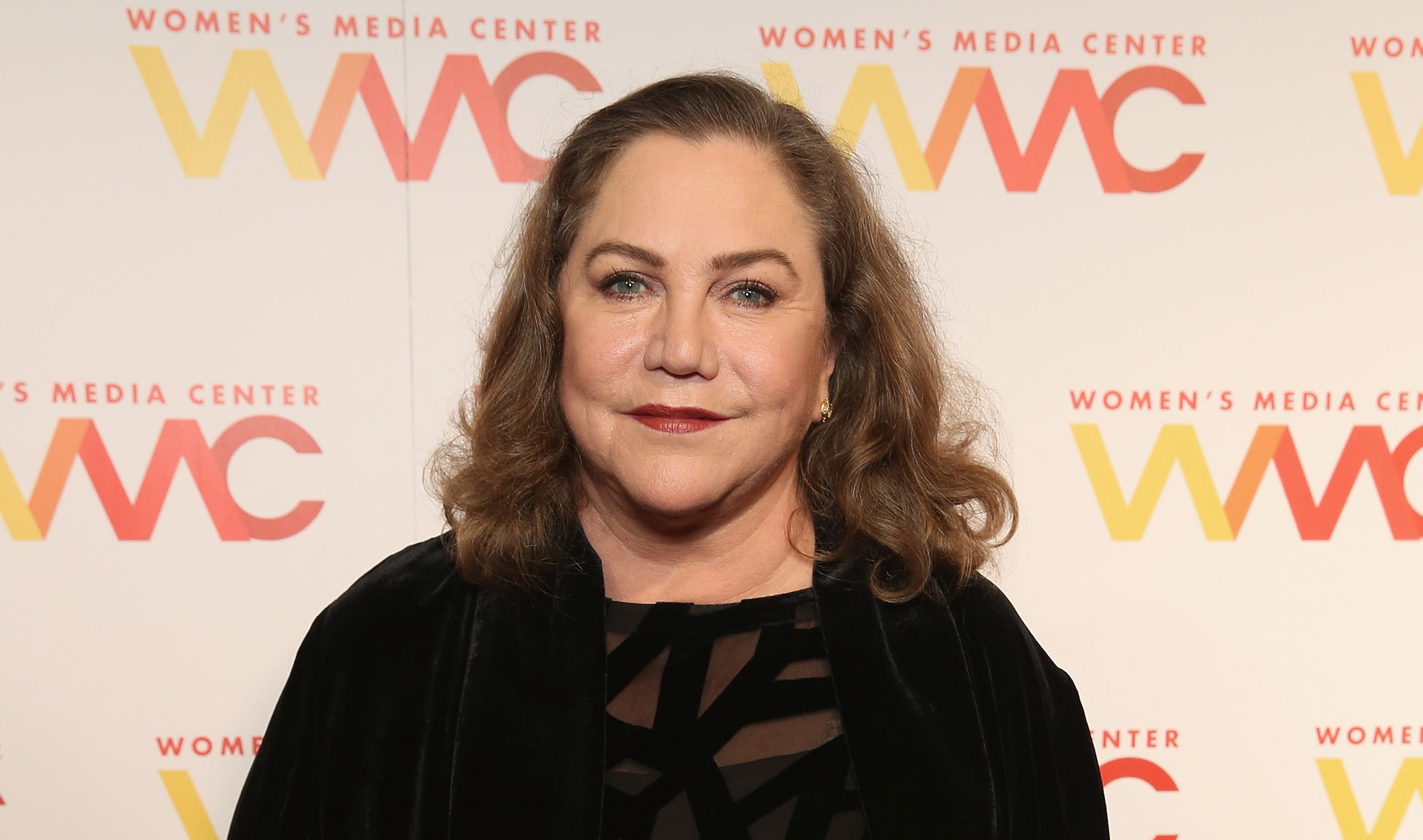 A photo shows actor Kathleen Turner wearing a black dress and smiling at the camera during a press event at the Women