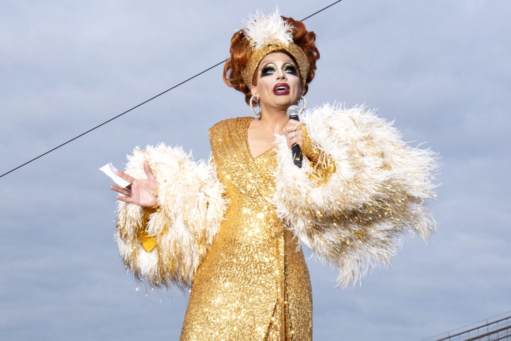 Biance Del Rio in a gold sequinned wrap dress and fur shrug, on the cusp of telling a joke