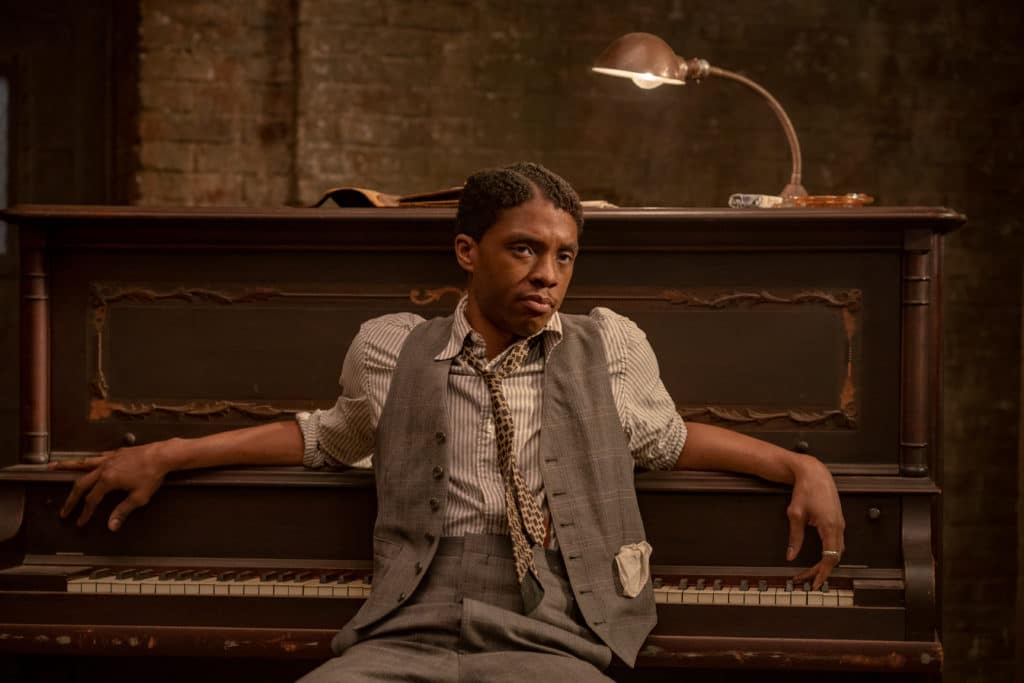 Chadwick Boseman sitting at a piano, facing away from the keys, in a grey waistcoat and low-slung tie