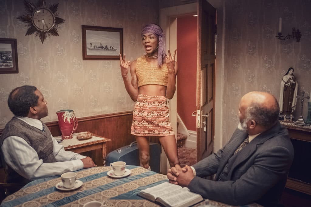 Roscoe in a skirt, crop top and head scarf, flipping the bird to his father and grandfather