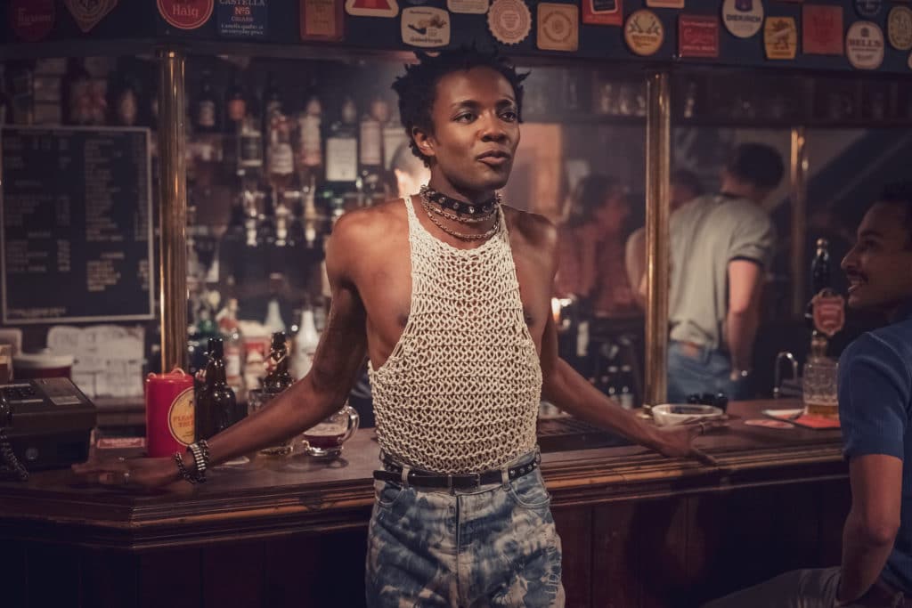 Omari Douglas as Roscoe, leaning against a pub bar, wearing acid-washed jeans and a string vest