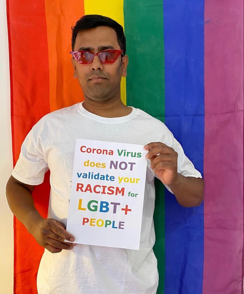 Nadim, a slim Bangladeshi man in a white t-shirt and sunglasses, standing in front of a rainbow flag holding a sign that says: &quot;Coronavirus does not validate your racism for LGBT+ people&quot;