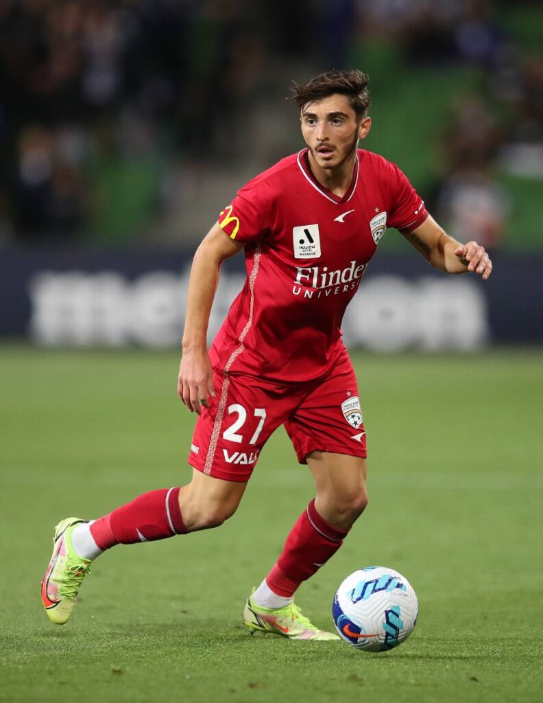 Josh Cavallo runs alongside a football while wearing a red jersey