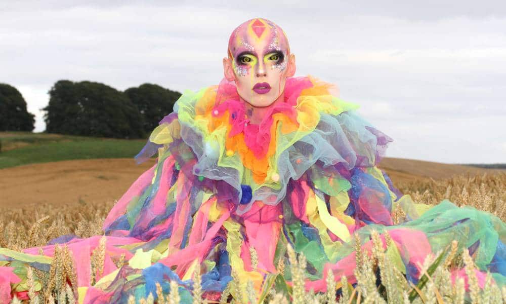 Cheddar Gorgeous, a drag artist, wears a rainbow coloured outfit made of different strips of tulle fabric with colourful makeup as they sit in field