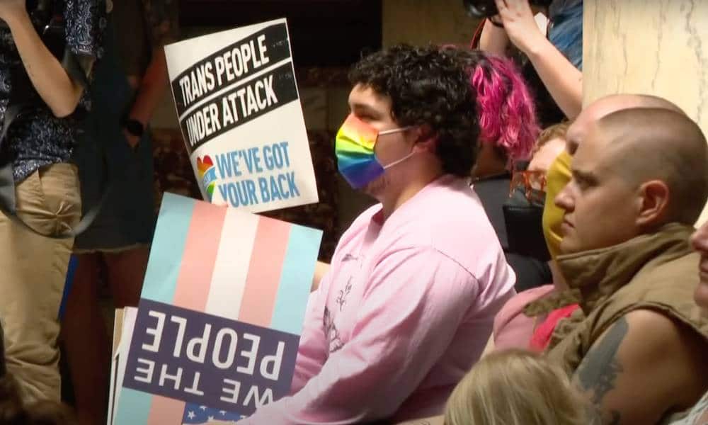 A person wearing a rainbow face mask holds a sign that in the colour of the trans flag while a sign reading 