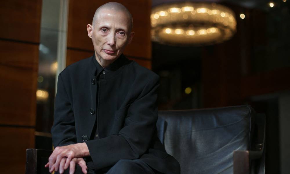 Christie Elan-Cane wears a dark black outfit with per hands crossed over each other while resting on per knee