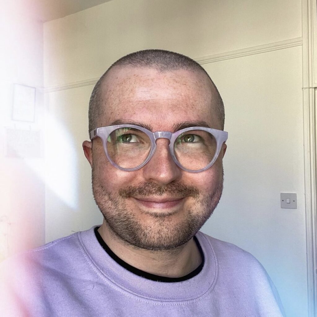 A picture of Tom Pashby: they have a closely-shaved head, are smiling and wearing white glasses