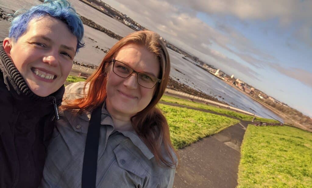 In this photograph, a person with blue hair wears a dark jacket as they embrace their partner with red hair and a grey jacket. 