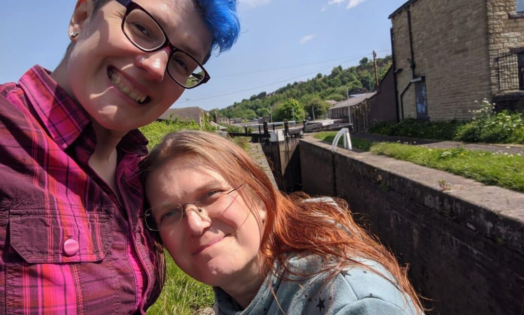 In this photograph, a couple are posed outside with one person, who has blue hair, wearing a red and purple plaid shirt. Their partner, who has red-brown hair, is posed near their chest wearing a grey top with stars on it
