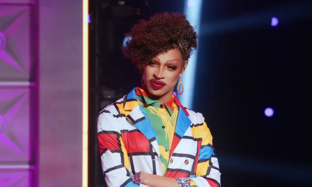 Drag Race star Yvie Oddly wears a white, blue, red and yellow geometric patterned outler layer with another colourful layer of clothing below. Yvie
