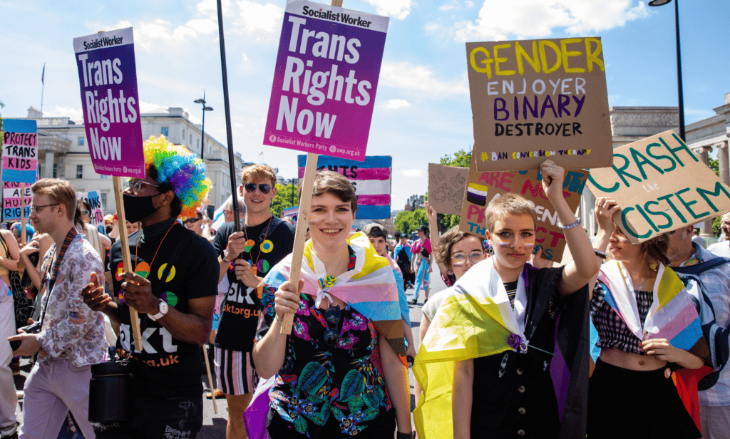 Several people gather in a crowd holding up signs in support of the trans community and LGBTQ+ people