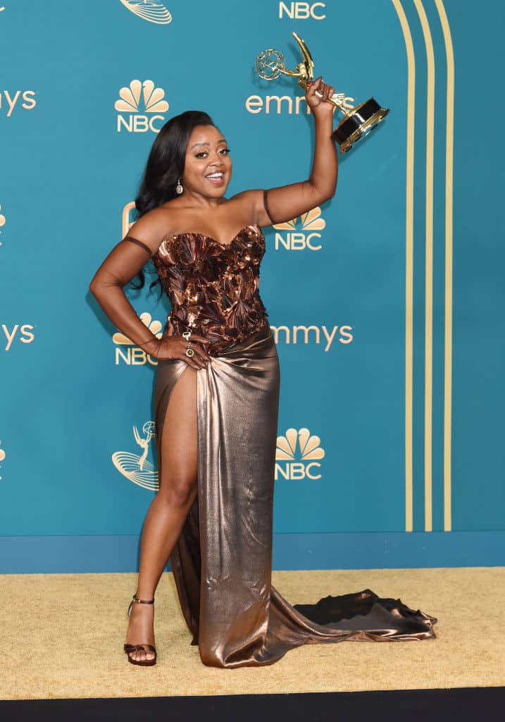 Quinta holding her Emmy aloft, wearing a metallic brown dress with embelissed embellished