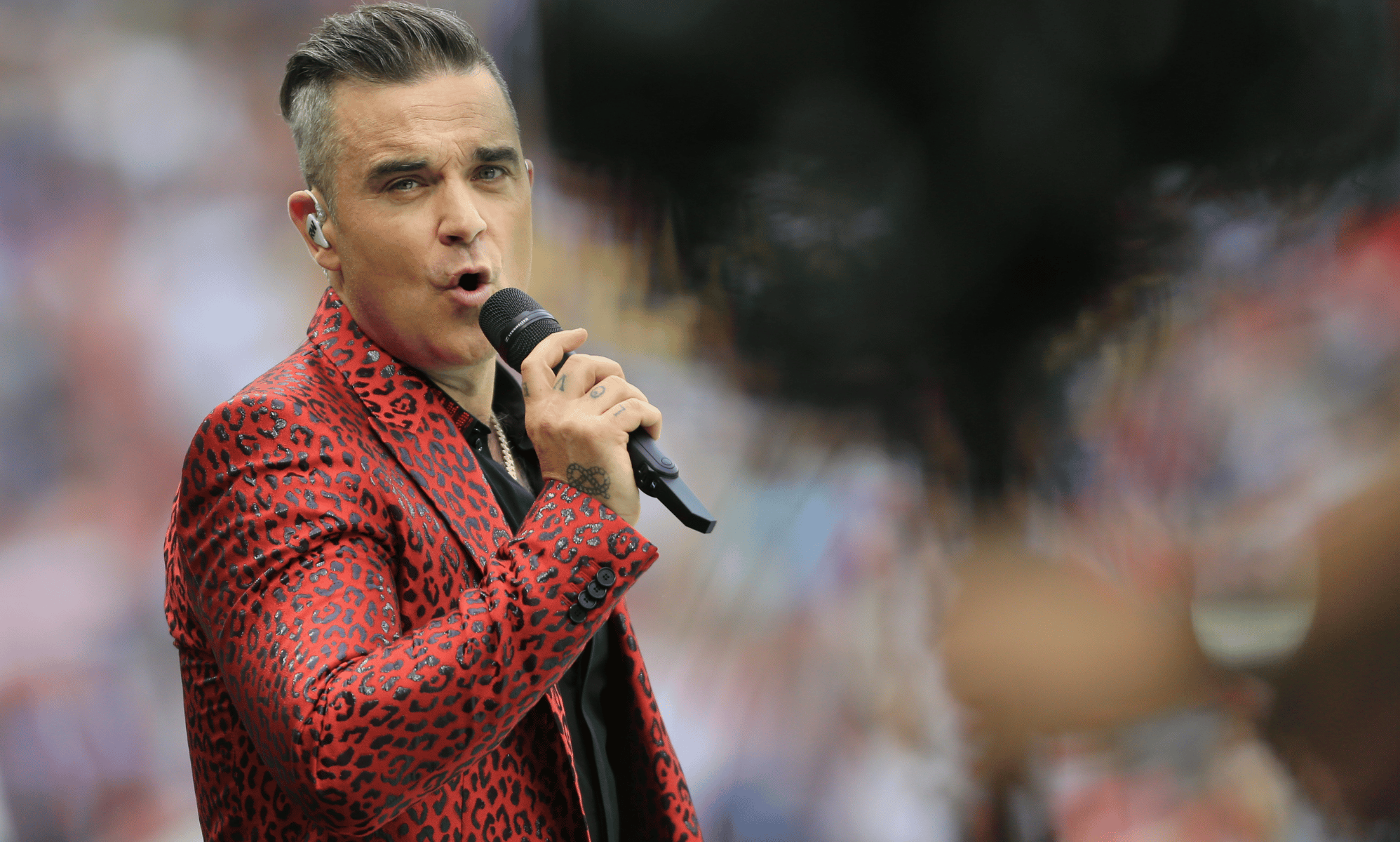 Robbie Williams wears a red animal-print patterned outfit as he sings into a microphone
