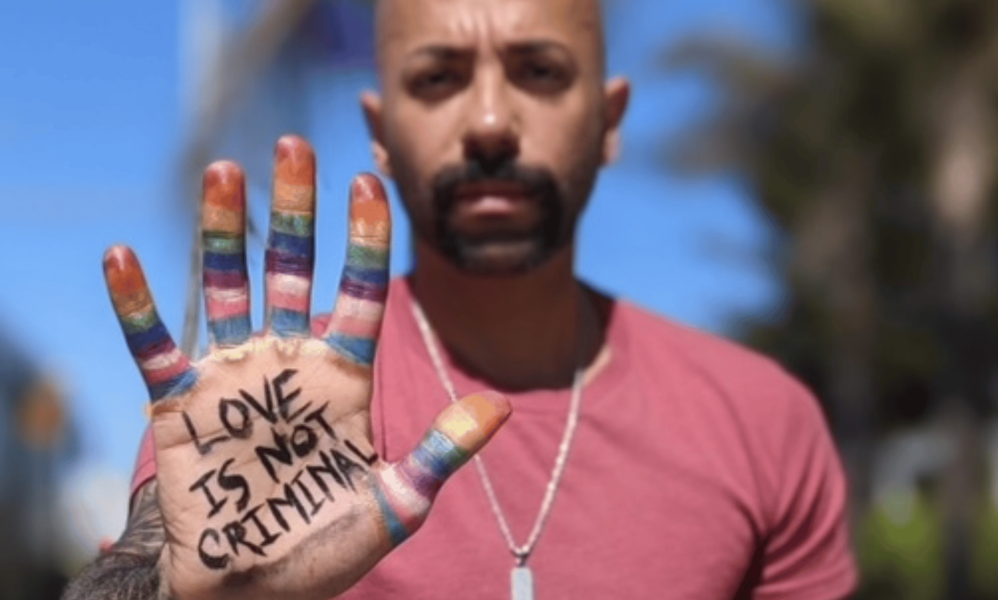 Dr Nas Mohamed, only publicly out LGBTQ+ Qatari, stands with his hand in front of him. His palm is facing towards the camera, and there are painted stripes in the colours of the LGBTQ+ and trans Pride flags on his fingers. In the centre of his palm, there are the worlds 