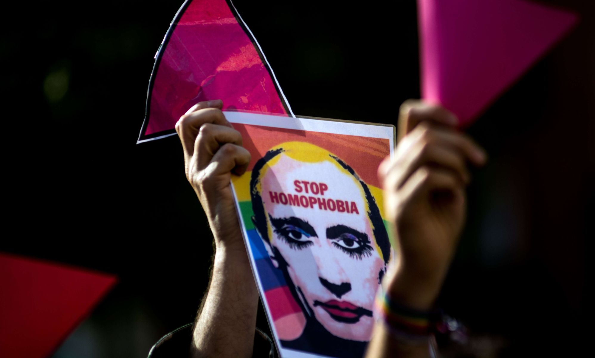 A person holds up a sign with a depiction of Russian president Vladimir Putin with LGBTQ+ rainbow colours in the background with the words 
