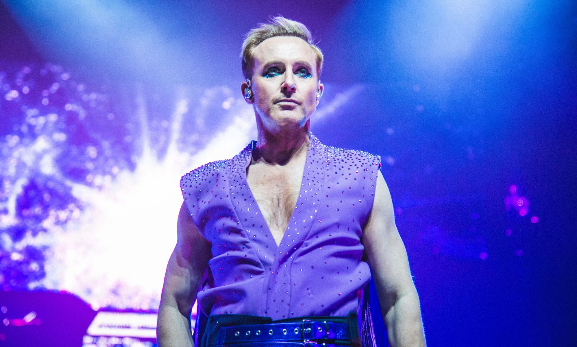 A photo of Ian &quot;H&quot; Watkins of Steps wearing a purple shirt with sequins as he stands on a stage with spotlights shining behind him