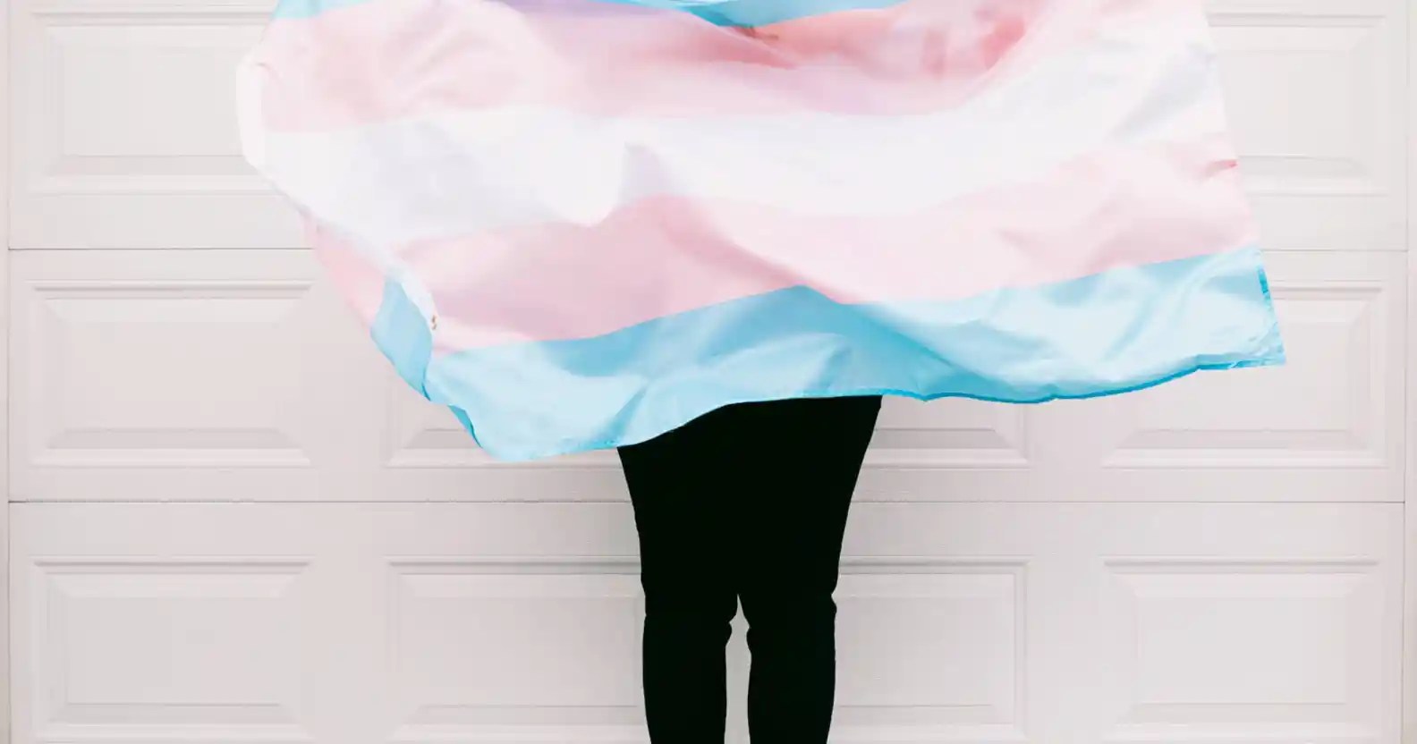 An image showing a person holding a transgender flag against a pale pink wall. The picture does not show the person