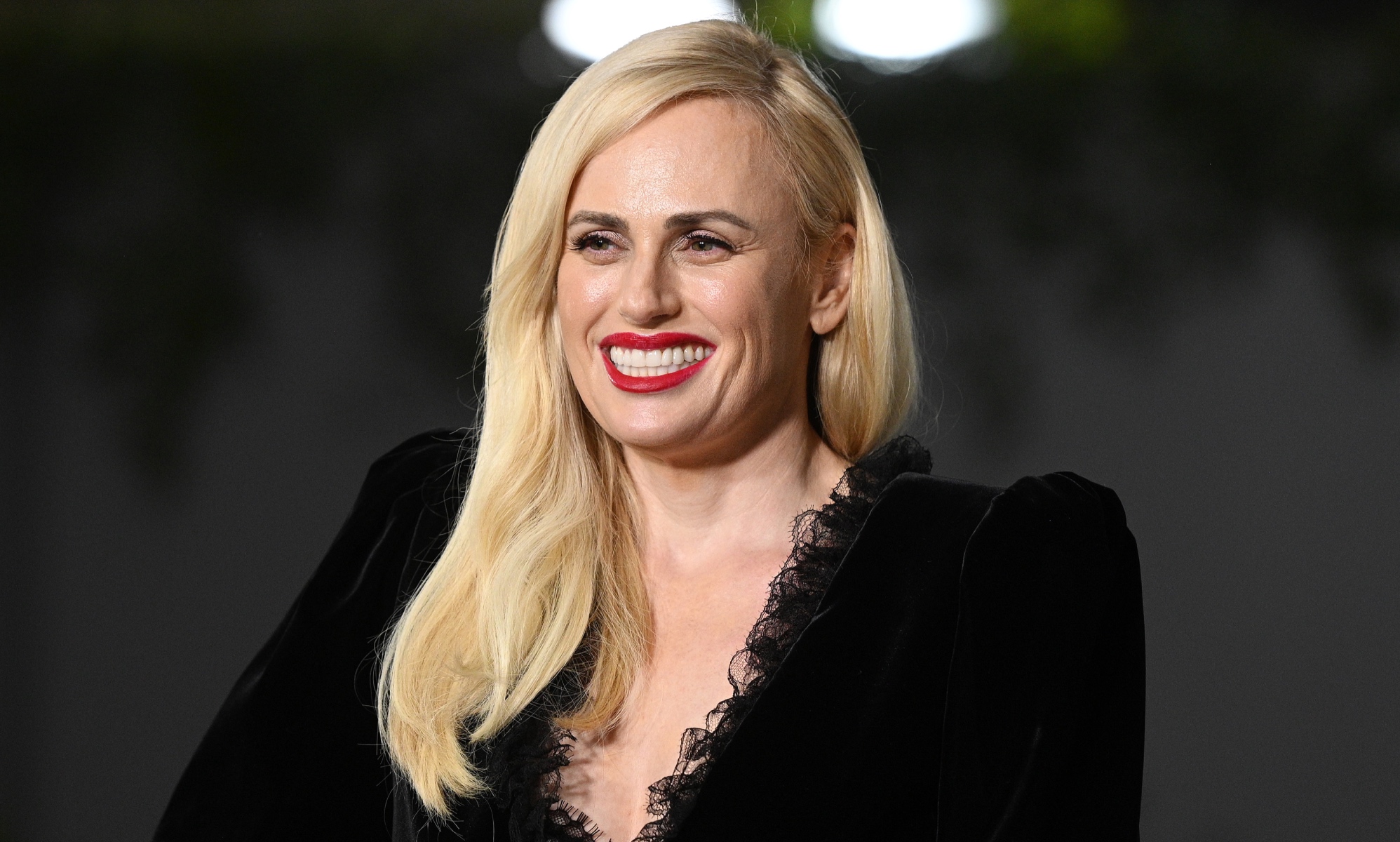 Photo of actor Rebel Wilson dressed in a black dress smiles for the cameras at a press event
