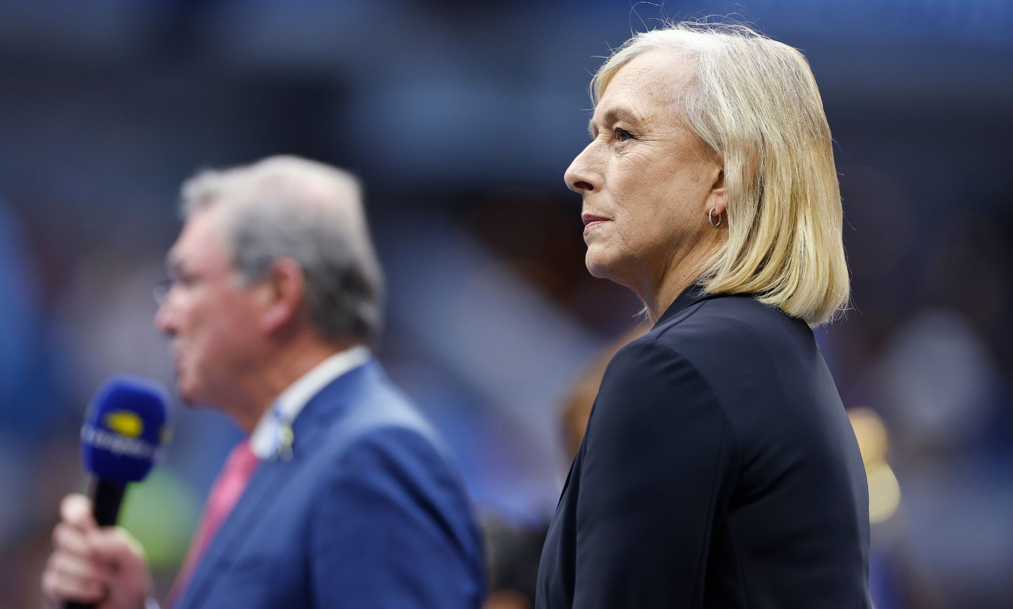 Martina Navratilova standing in a Tennis court, next to an individual wearing a blue blazer and holding a microphone.