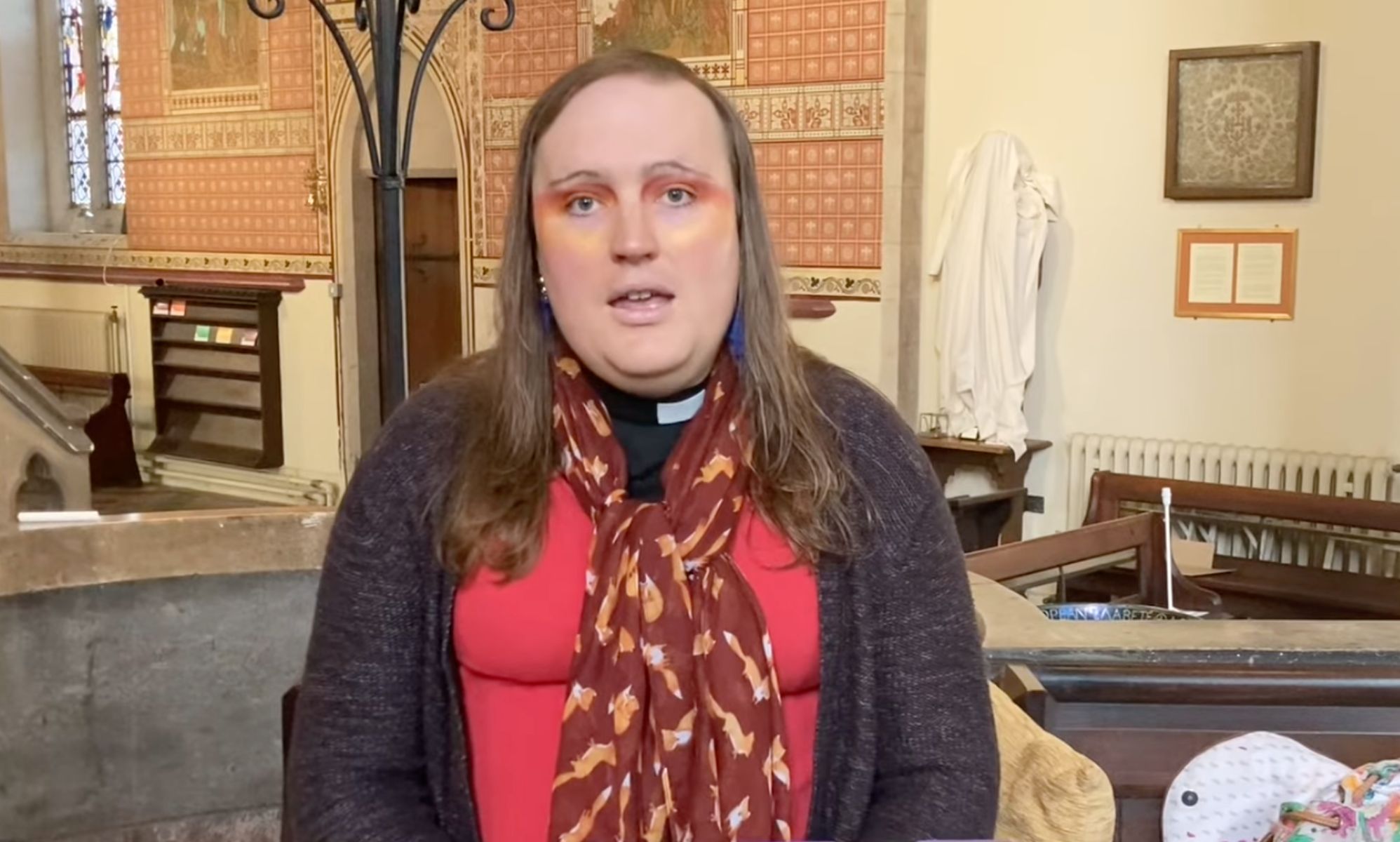 Bingo Allison, a non-binary, genderqueer Church of England priest, wears a red top, sweater and scarf while they speak about religion and Trans Day of Visibility