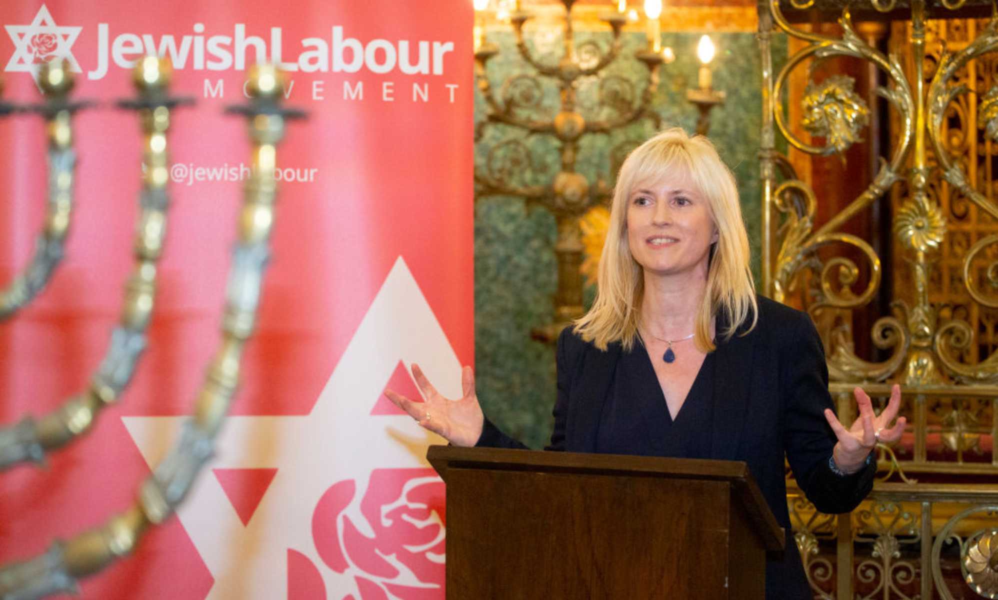 Roșie Duffield speaking at a Jewish Labour event in 2019. Duffield is pictured on the right hand side of the image standing at a podium wearing a black suit. On the left is Jewish Labour signage.