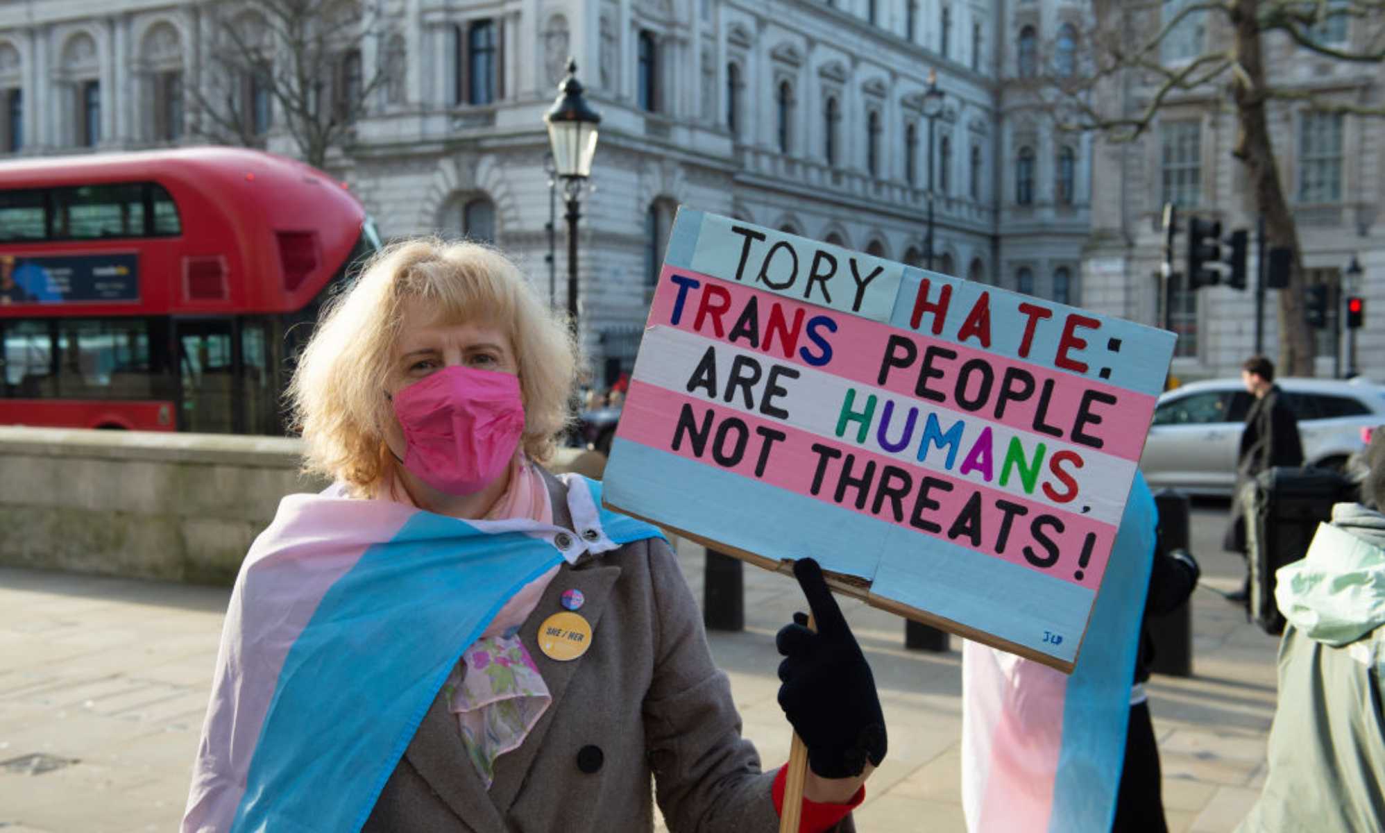 A trans activist takes part in a protest opposite Downing Street on 17 January 2023. The person is holding a sign that reads: &quot;Tory hate: Trans people are humans not threats!&quot; the sign is coloured in the colours of the trans flag, and the person is wearing a trans Pride flag draped around them.