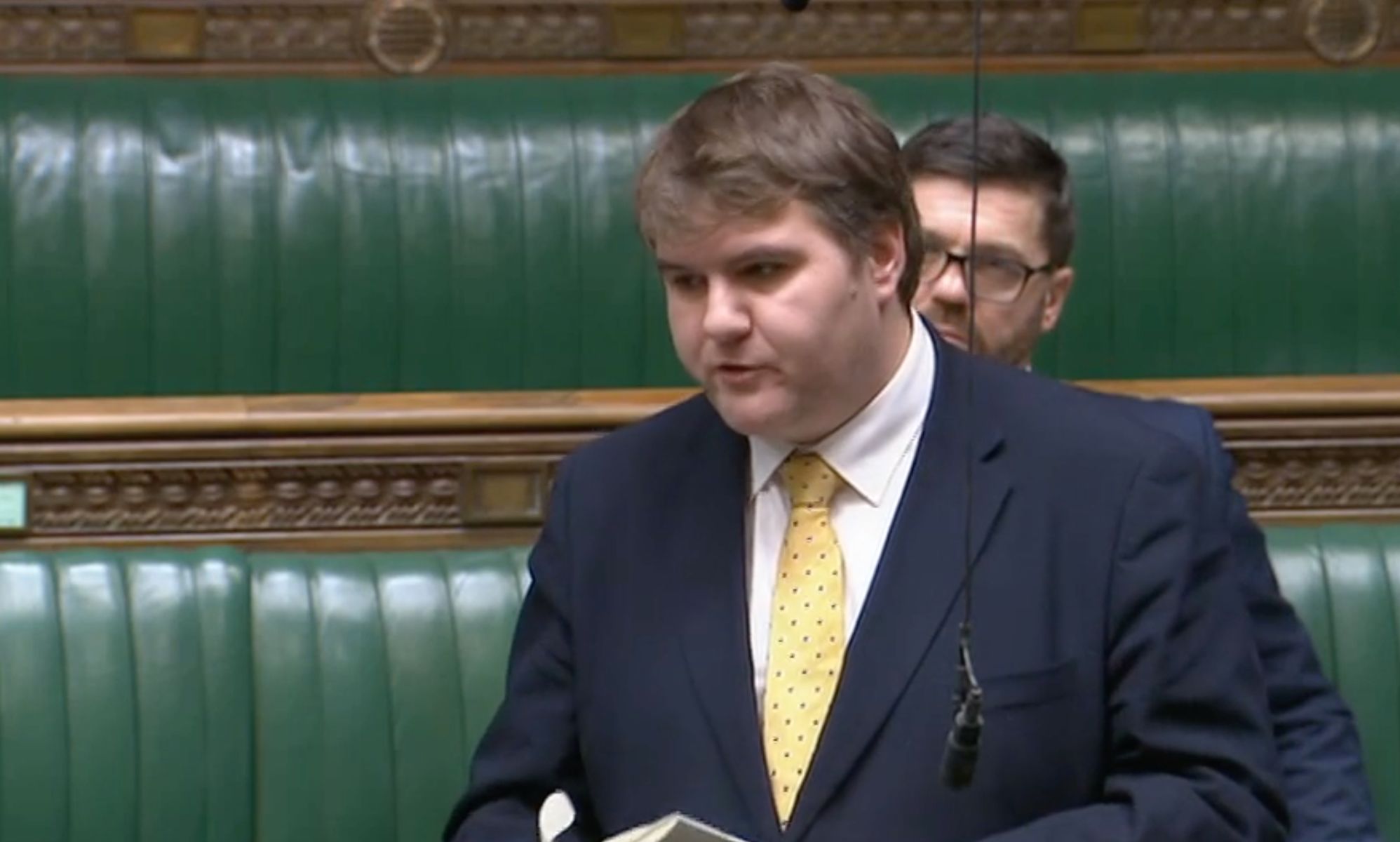 Jamie Wallis wears a white shirt, yellow tie and a blue jacket while addressing parliament