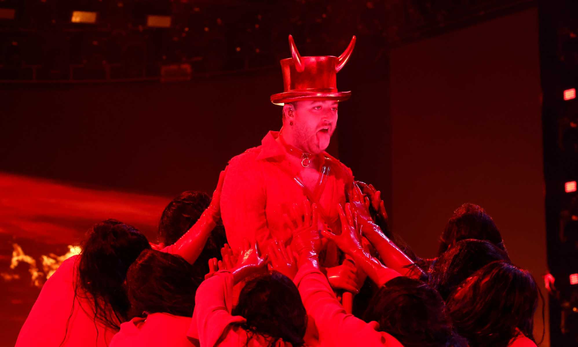 A screenshot from the 2023 Grammys ceremony shows Sam Smith performing &quot;Unholy&quot; on stage wearing a hat showing devil horns as a red spotlight casts a strong red light over him