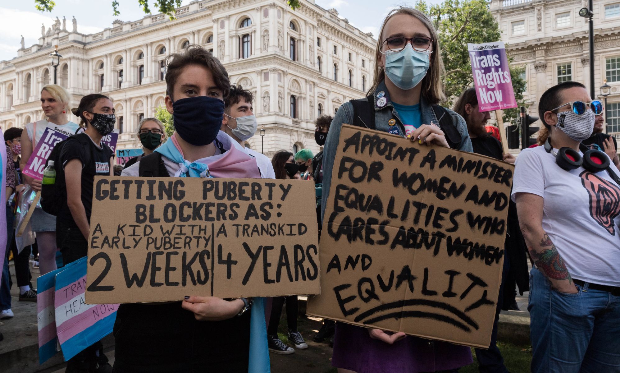 Two people are pictured holding up signs in support of the trans community at a protest. One person