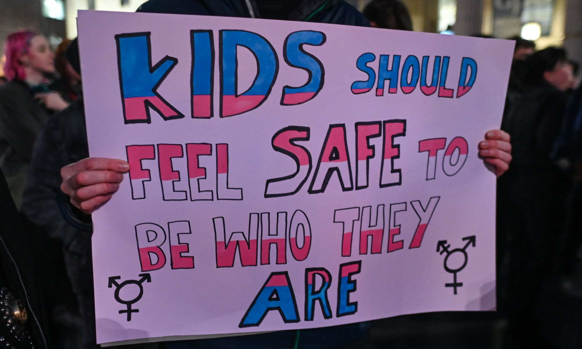 A person holds a sign reading &quot;kids should feel safe to be who they are&quot; in the colour of the trans pride flag