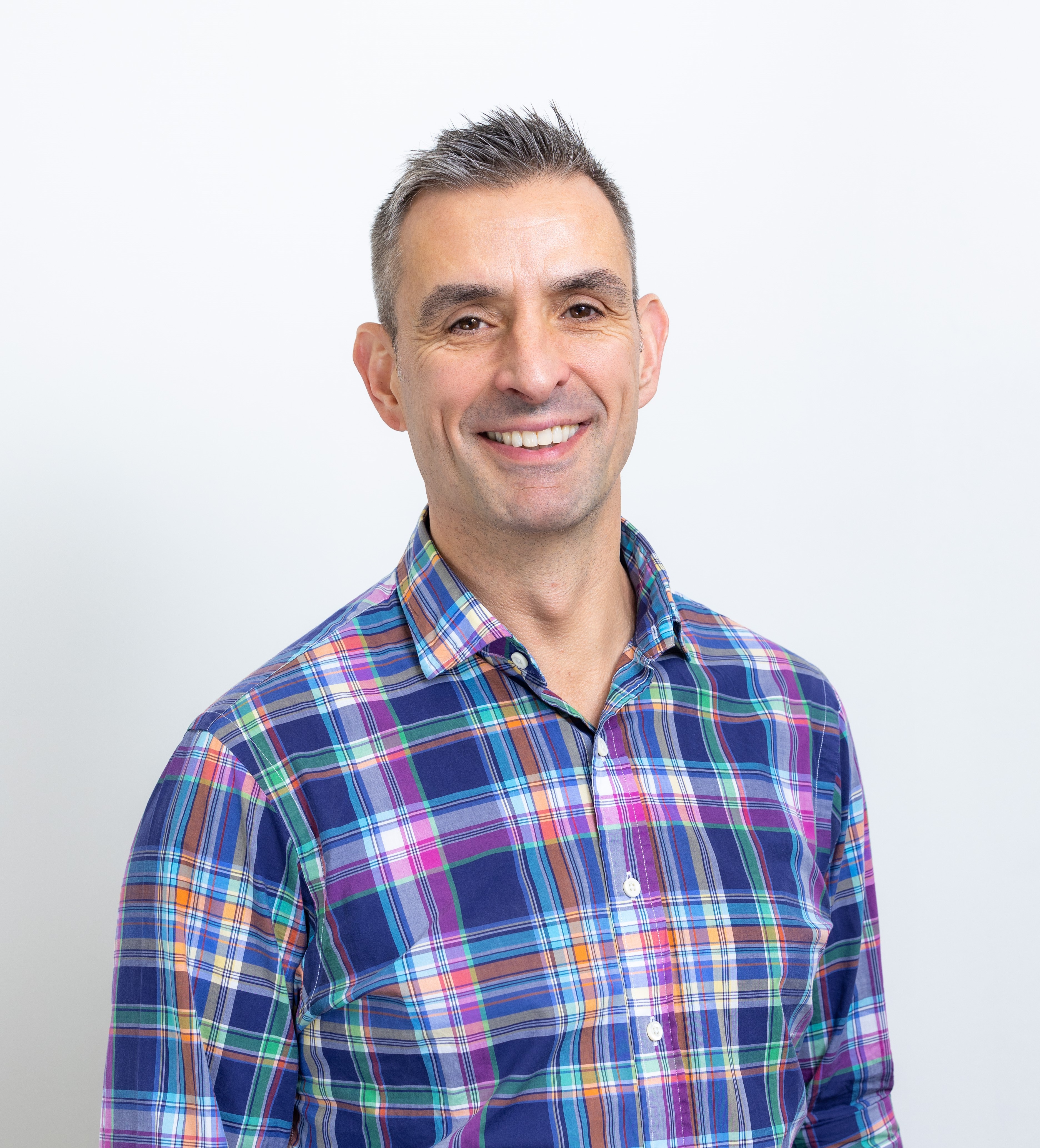 Simon Blake is wearing a plaid shirt and is smiling at the camera.