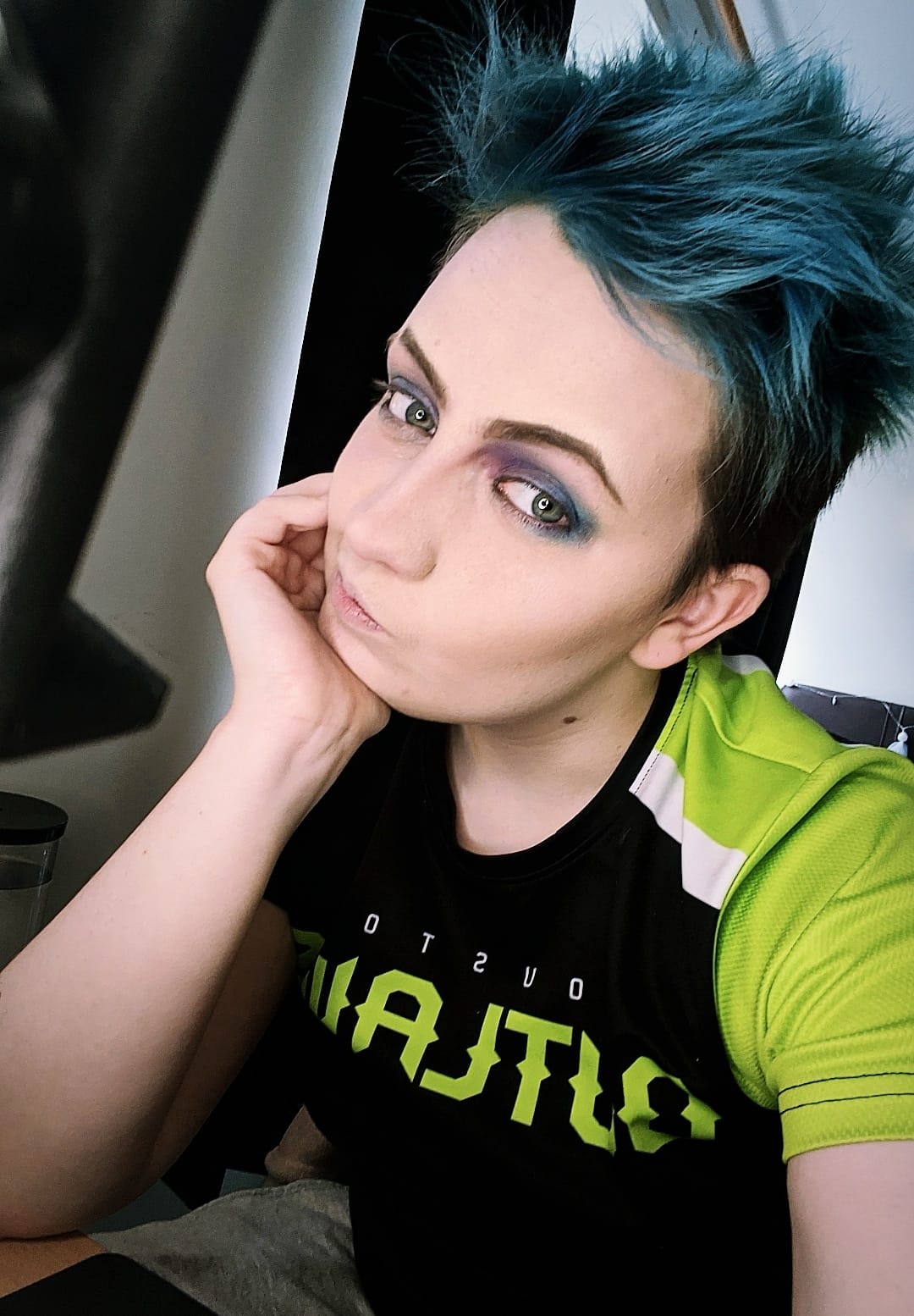 Maxie is taking a selfie. They have short blue hair and has their face resting on their hands.