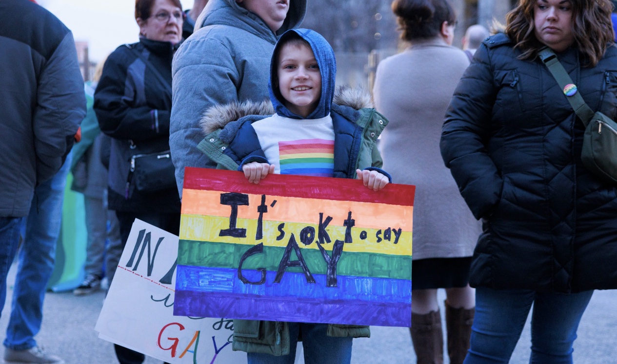 A child in a rainbow T-shirt holds a sign reading &quot;It