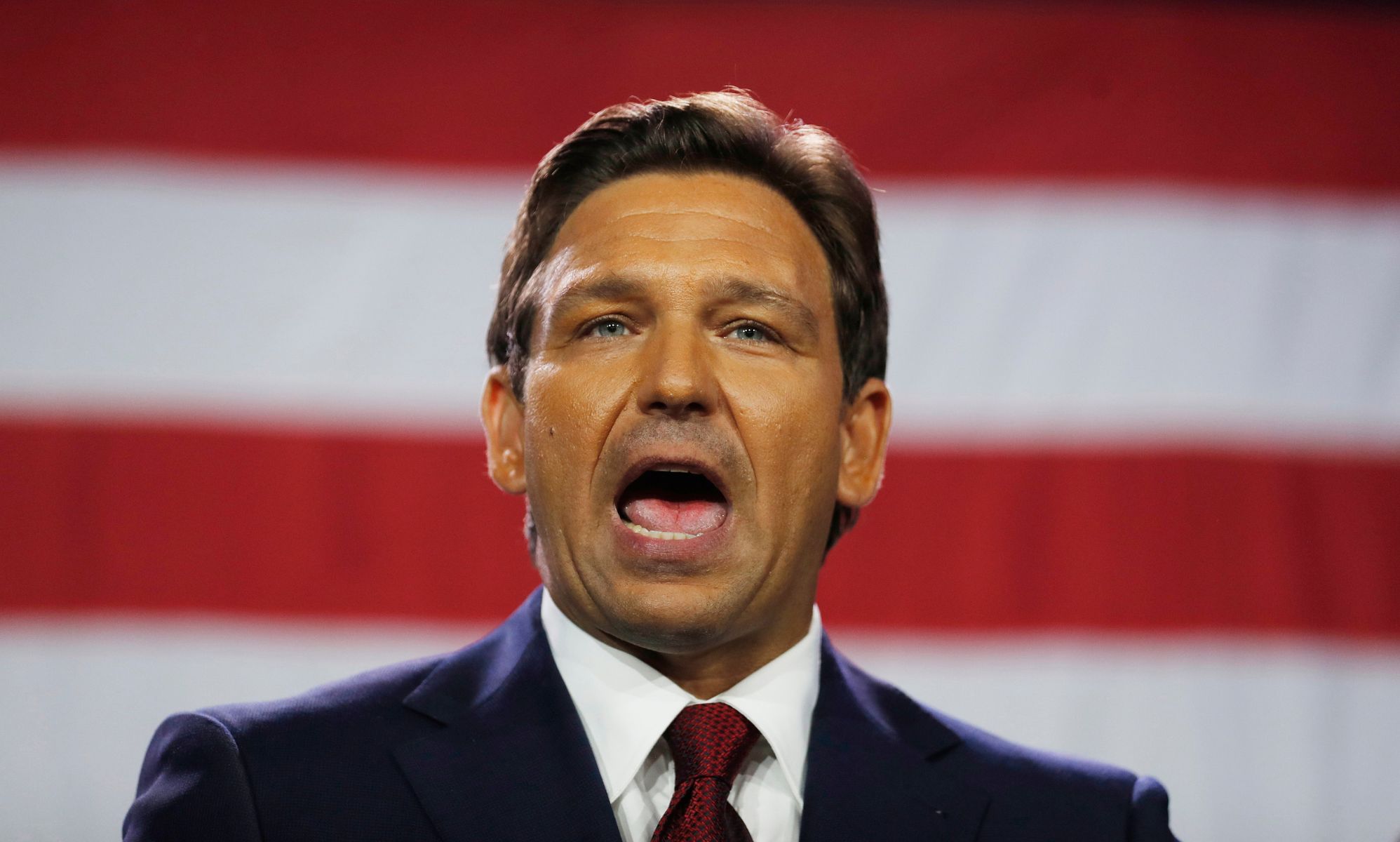 Florida governor Ron DeSantis wears a suit and tie as he speaks with his mouth wide open. The red and white stripes of the US flag can be seen in the background