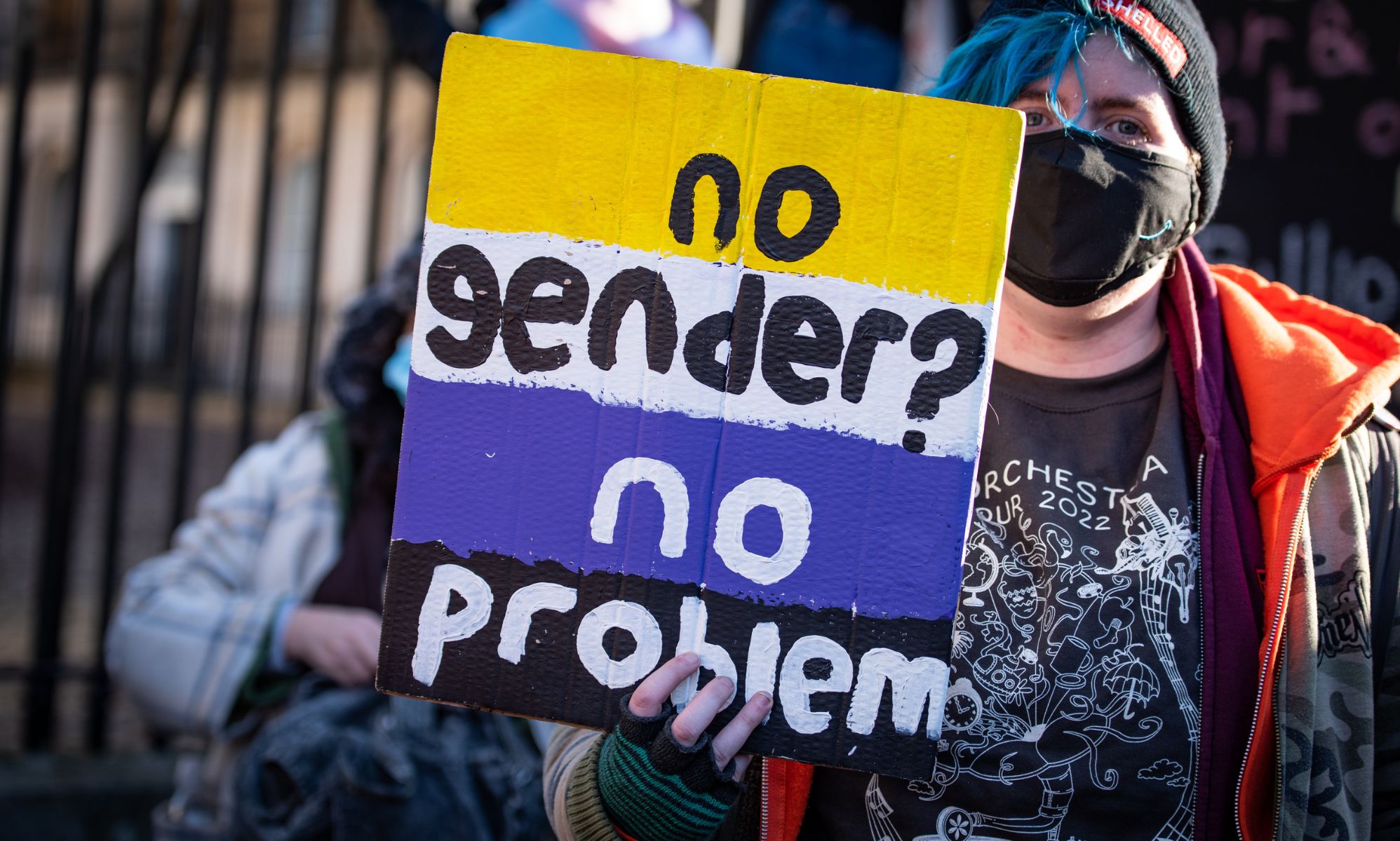 A person holds up a sign in the colours of the non-binary flag (yellow, white, purple and black) with the words &quot;No gender? No problem&quot;