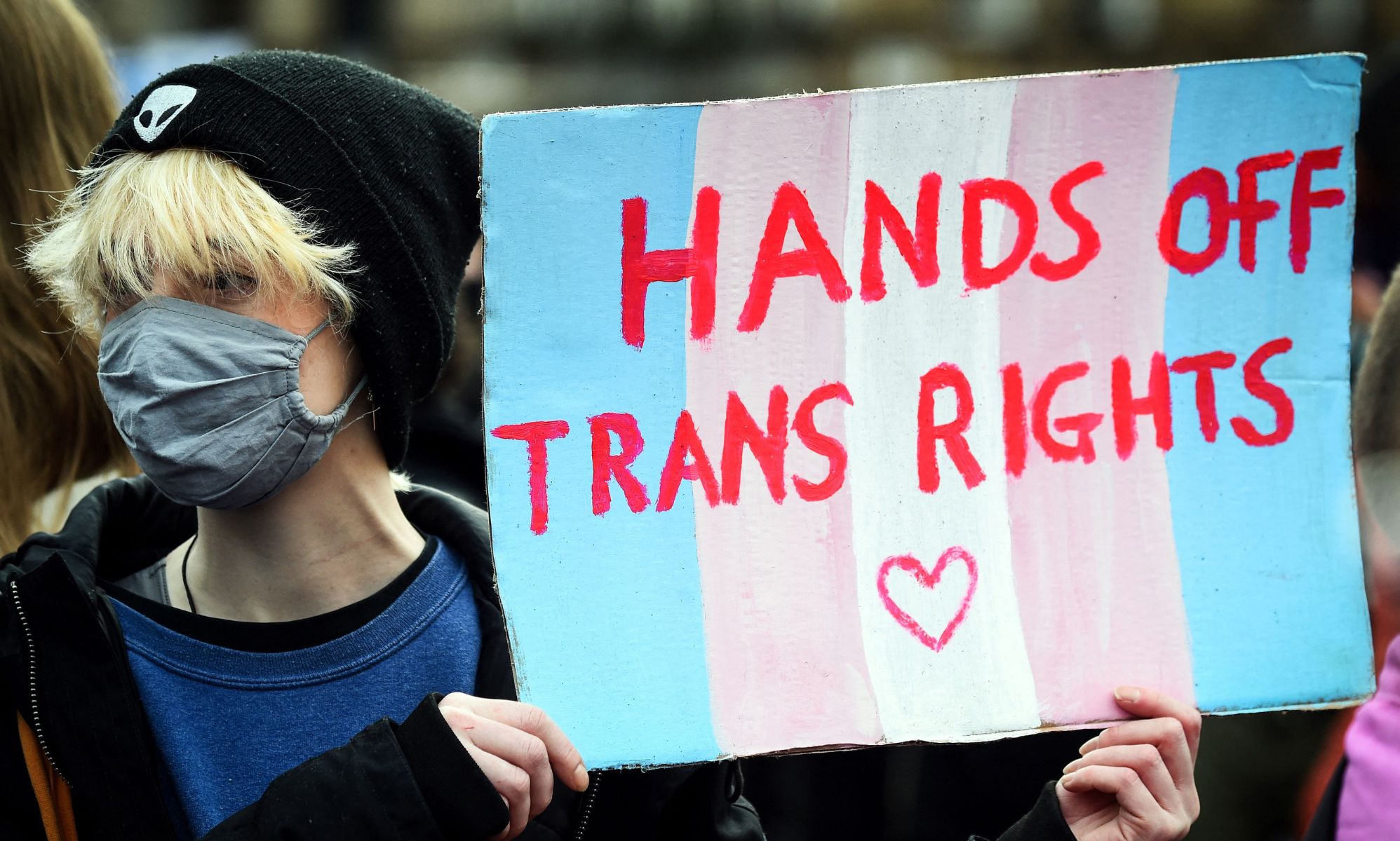 A person holds up a sign in the colours of the trans pride flag (blue, pink and white) reading 