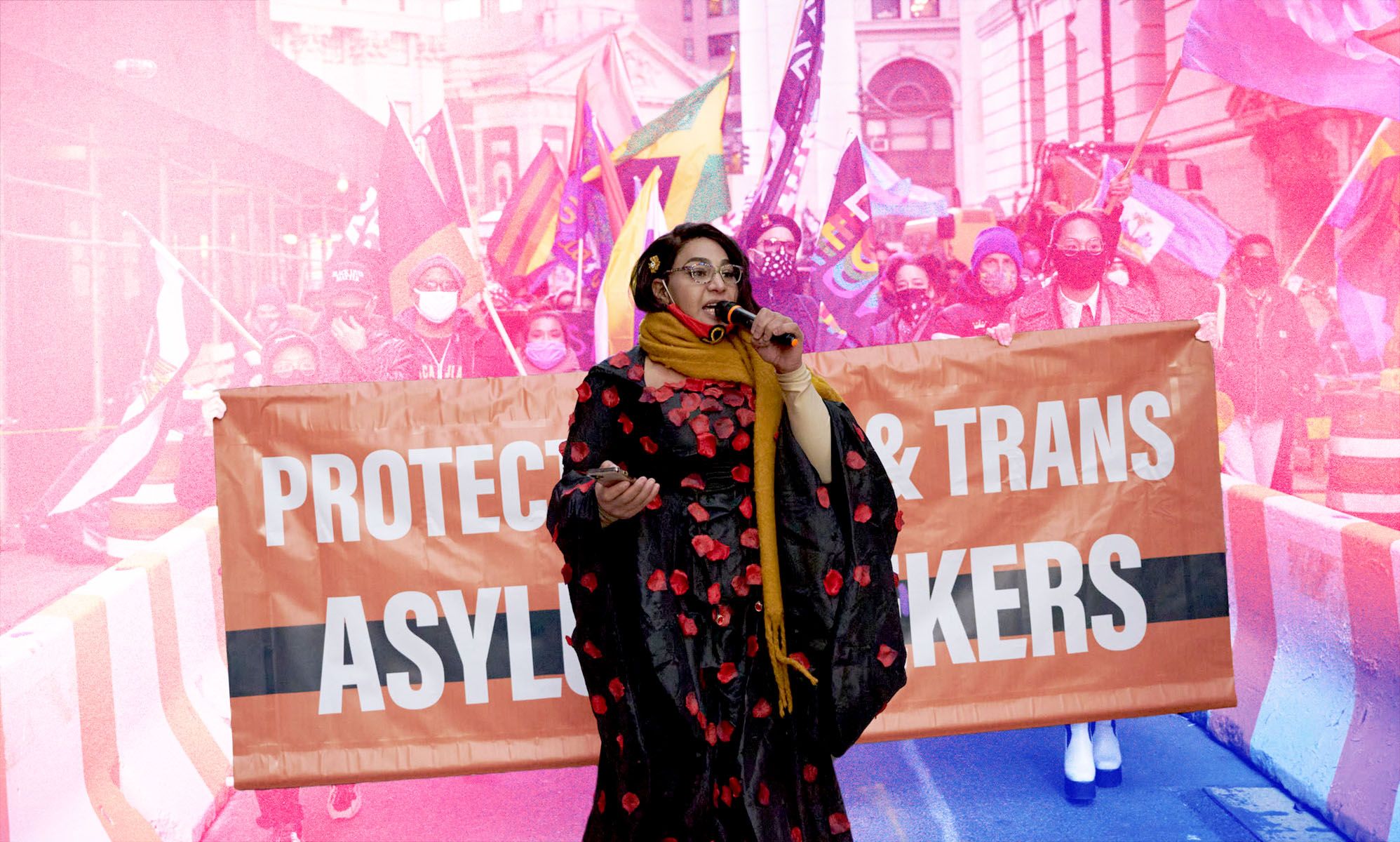 Trans activist Iman Le Caire speaks passionately into a microphone as she stands in front of a banner depicting its support for asylum seekers amid a march with a pink and blue wash over parts of the image
