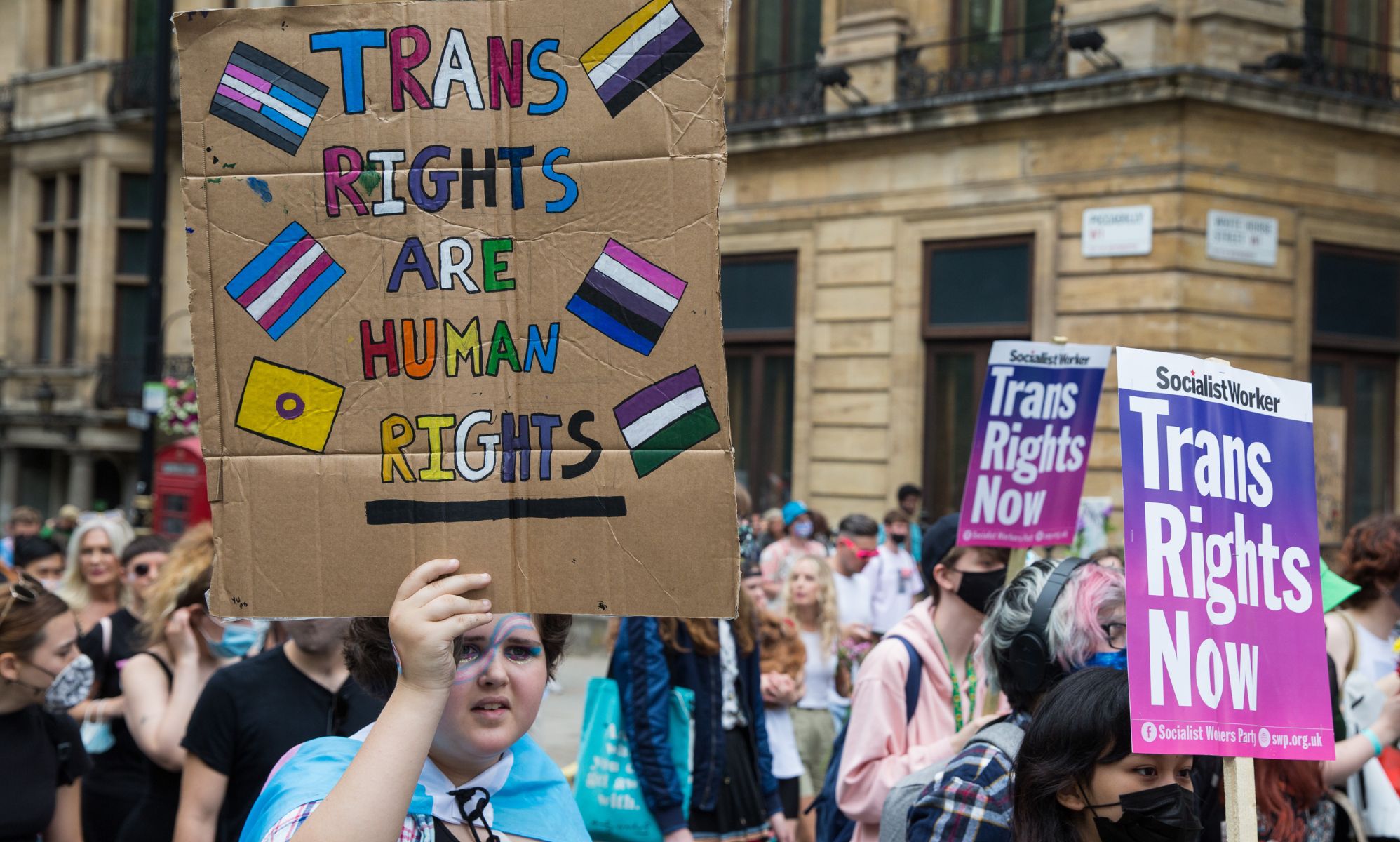 A person holds up a sign with the non-binary, trans, intersex, genderqueer, genderfluid flags on it reading 