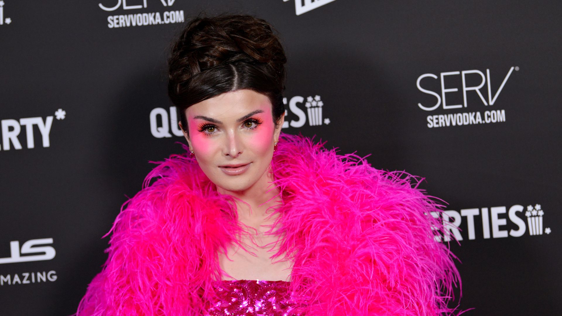 Dylan Mulvaney wears pink feather boa and pink eyeshadow against a black background