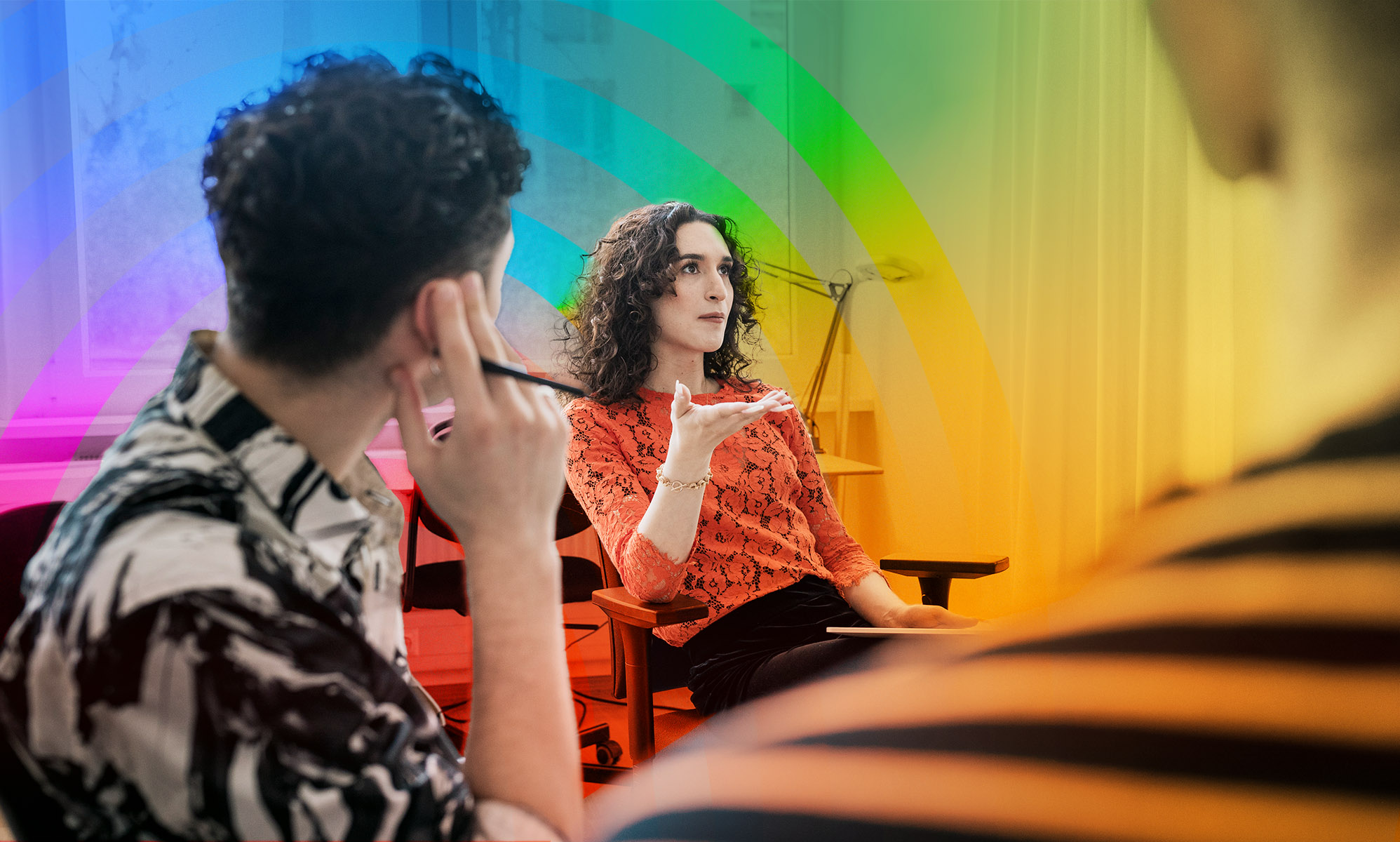 In this picture, a trans woman is speaking to a group of people. She is wearing orange and has curly brown hair. 