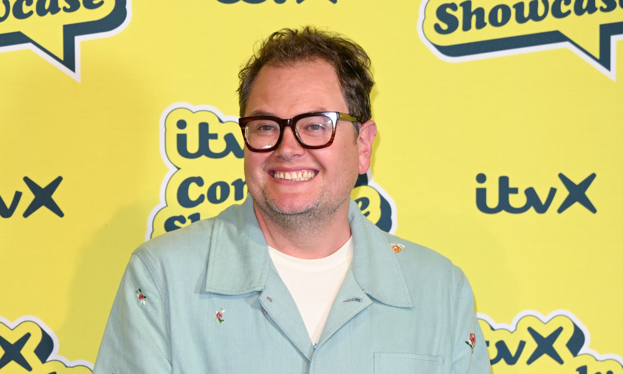 Alan Carr in a mint green shirt and white t-shirt smiling and standing in front of a yellow background.
