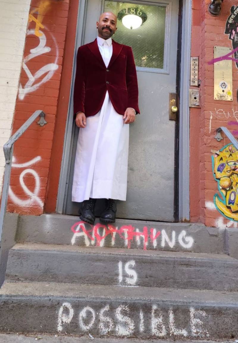 LGBTQ+ rights activist Dr Nasser Mohamed, who is from Qatar, wears a white outfit as he stands on the steps to a building with the words 