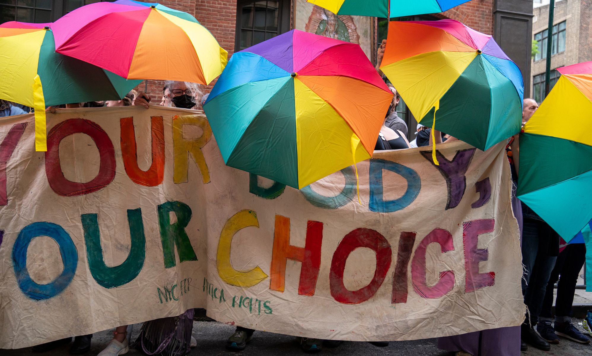 A crowd of people hold up rainbow coloured umbrellas with a sign reading 
