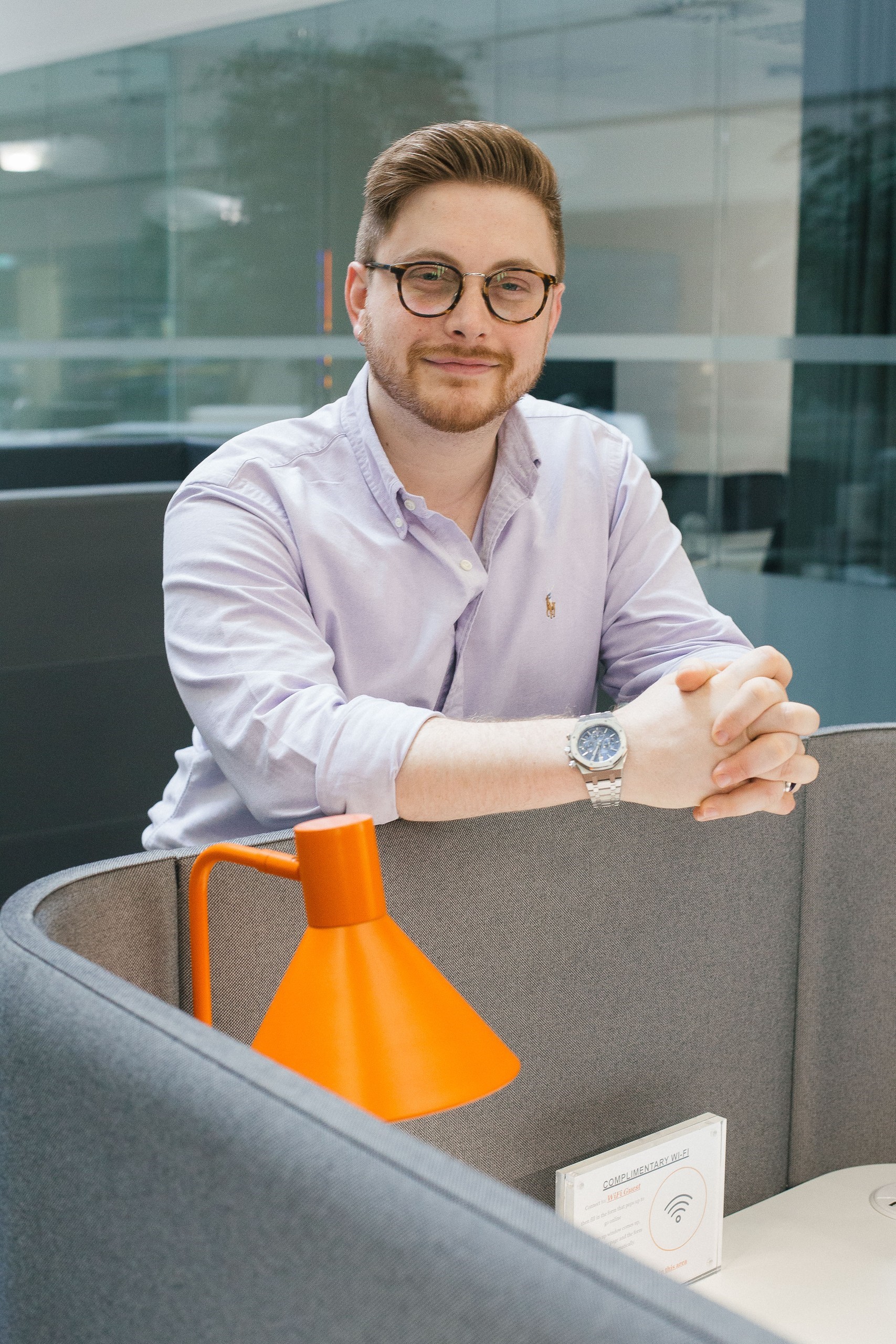This is an image of Joseph Williams, he is standing in an office cubilce. He has reddish hair and is wearing a light coloured buttoned shirt and dark rimmed rounded eyeglasses. His hands are folded on the workspace divider.