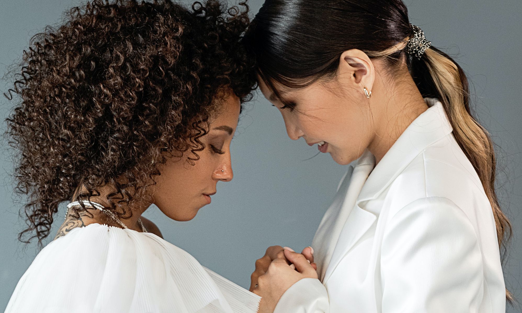 A queer, same-sex couple wear white outfits as they attend a wedding and hold each other