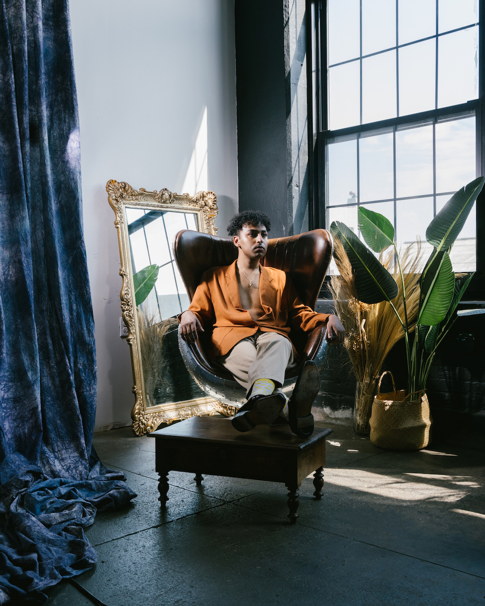 Rufus Sivaroshan, a music artist, pictured in a posed image sitting on an armchair with a mirror behind them and a decorated room in the background.