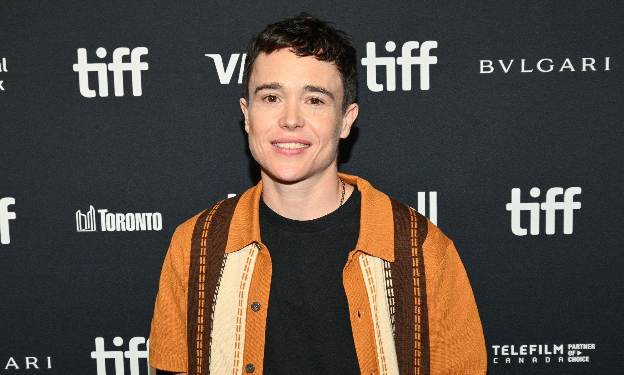 Elliot Page smiles on the Toronto Film Festival red carpet. He is wearing a black top and orange and brown shirt.