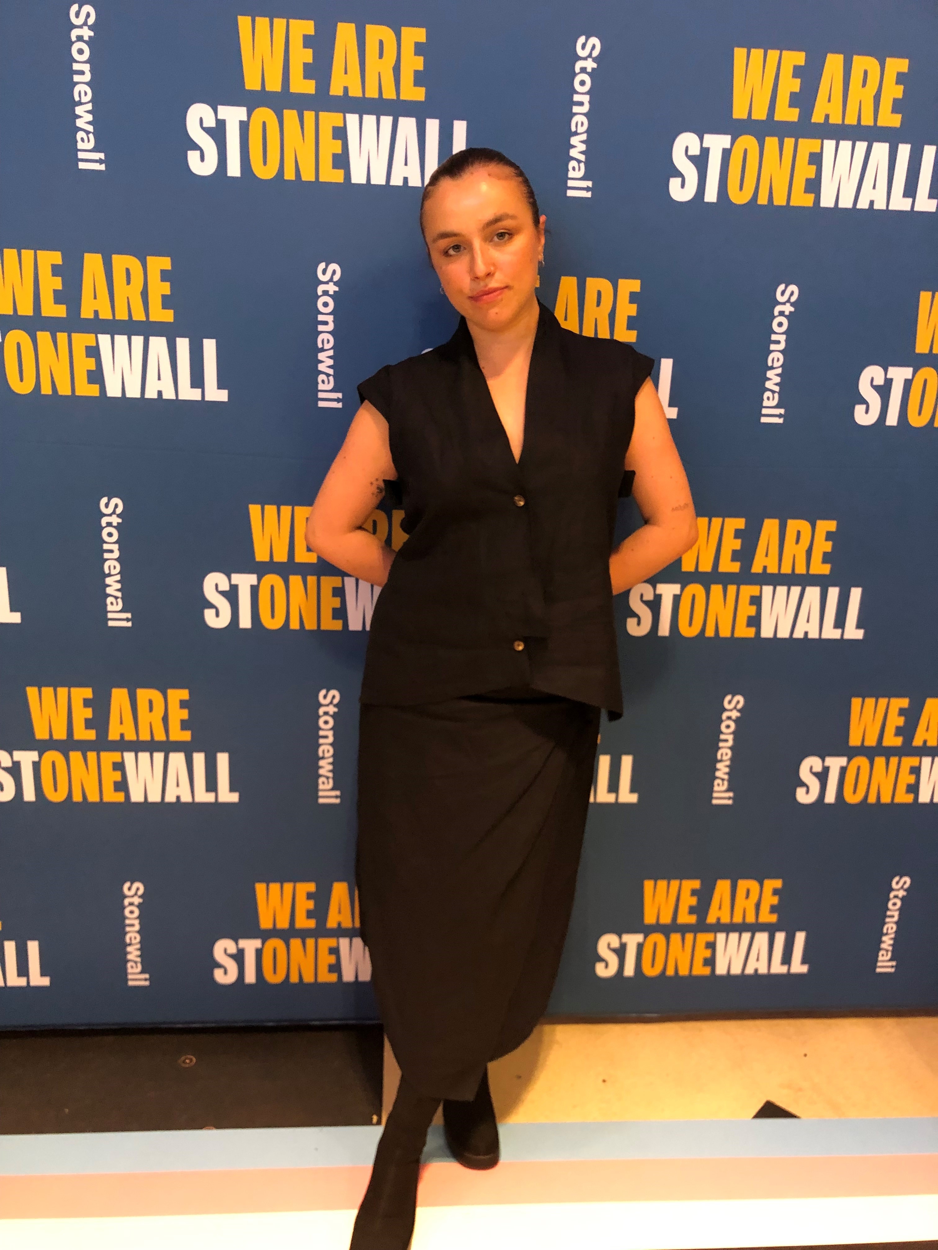 This is an image of a woman stand on a blue backdrop with the words &quot;We are Stonewall&quot; in yellow and white. She is wearing a black dress.